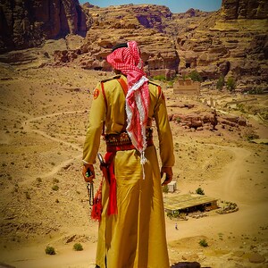 Puede incluir: Un hombre con un keffiyeh rojo y blanco tradicional se encuentra en un paisaje desértico, mirando hacia el terreno rocoso. Lleva una túnica de color canela y un cinturón con una funda. El cielo es azul y el sol brilla.