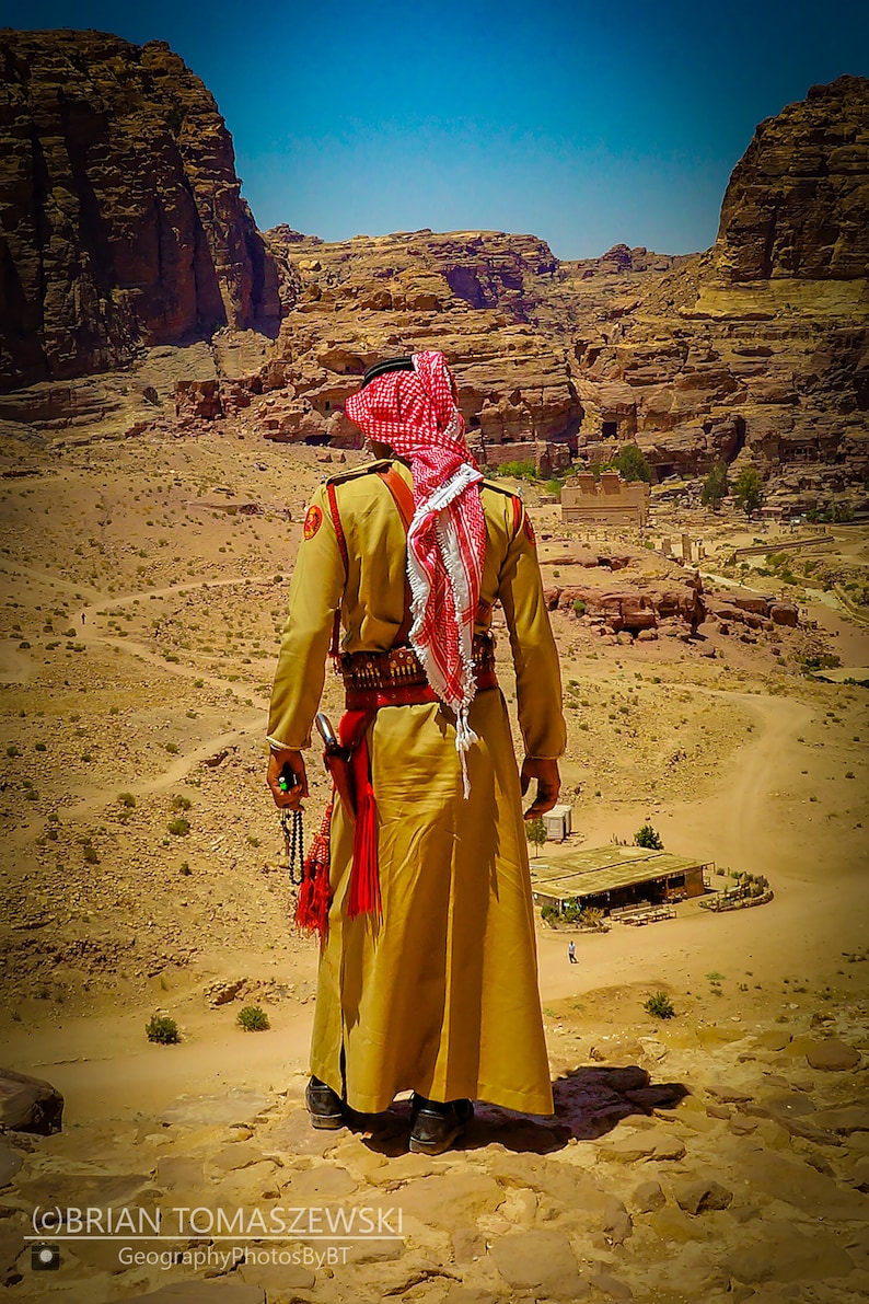 Puede incluir: Un hombre con un keffiyeh rojo y blanco tradicional se encuentra en un paisaje des&eacute;rtico, mirando hacia el terreno rocoso. Lleva una t&uacute;nica de color canela y un cintur&oacute;n con una funda. El cielo es azul y el sol brilla.