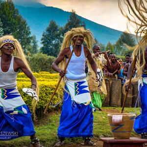 May include: A group of people in traditional clothing are dancing and holding sticks. They are wearing blue and white skirts and have long, flowing hair. The background is a green field with trees and mountains in the distance.