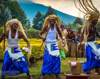 Puede incluir: Un grupo de personas con vestimenta tradicional están bailando y sosteniendo palos. Llevan faldas azules y blancas y tienen el pelo largo y suelto. El fondo es un campo verde con árboles y montañas en la distancia.