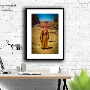 Puede incluir: Una fotograf&iacute;a enmarcada de una persona vestida con una t&uacute;nica tradicional y un pa&ntilde;uelo rojo caminando por un paisaje des&eacute;rtico. La foto est&aacute; enmarcada en negro y est&aacute; colgada en una pared de ladrillo blanco.