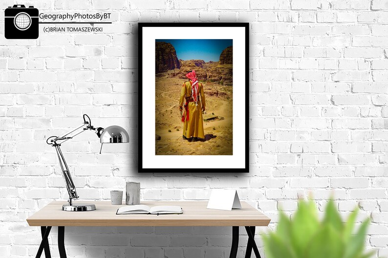 Puede incluir: Una fotograf&iacute;a enmarcada de una persona vestida con una t&uacute;nica tradicional y un pa&ntilde;uelo rojo caminando por un paisaje des&eacute;rtico. La foto est&aacute; enmarcada en negro y est&aacute; colgada en una pared de ladrillo blanco.