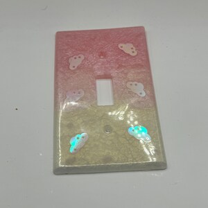 Puede incluir: Una tapa de interruptor de luz con un dise&ntilde;o degradado rosa y blanco y nubes iridiscentes con estrellas.