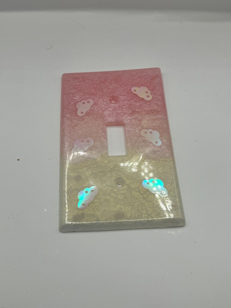 Puede incluir: Una tapa de interruptor de luz con un dise&ntilde;o degradado rosa y blanco y nubes iridiscentes con estrellas.