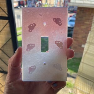 Puede incluir: Una tapa de interruptor de luz blanca y rosa con un dise&ntilde;o degradado. La tapa presenta un patr&oacute;n repetitivo de nubes blancas con peque&ntilde;as estrellas sobre un fondo rosa. La tapa del interruptor est&aacute; hecha de un material liso y brillante.