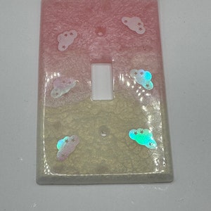 Puede incluir: Una tapa de interruptor de luz de color rosa claro y amarillo con un dise&ntilde;o de nubes y estrellas iridiscentes.
