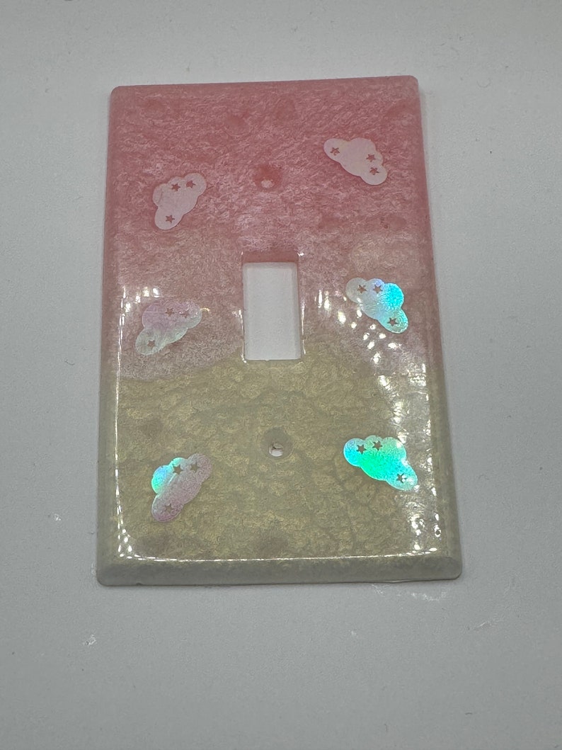 Puede incluir: Una tapa de interruptor de luz de color rosa claro y amarillo con un dise&ntilde;o de nubes y estrellas iridiscentes.