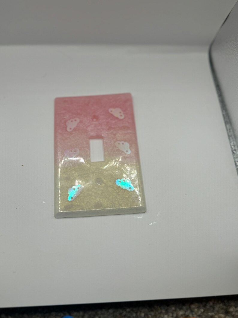 Puede incluir: Una tapa de interruptor de luz con un dise&ntilde;o degradado rosa y blanco y detalles de nubes iridiscentes.