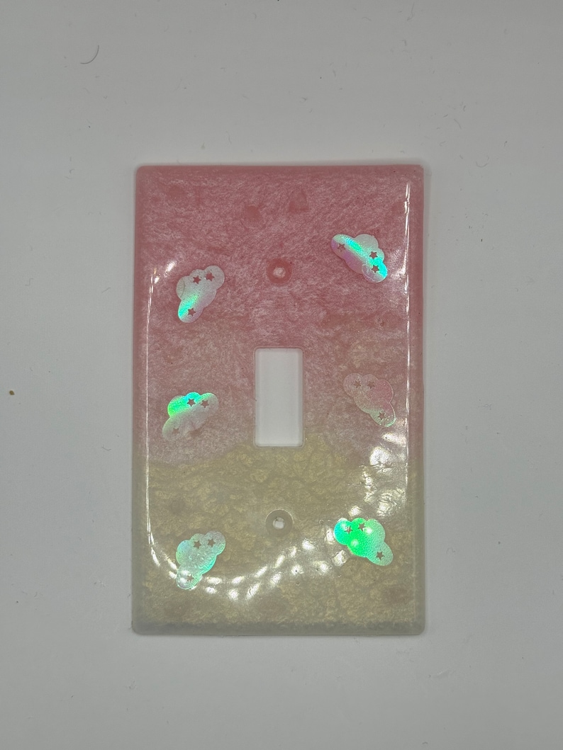 Puede incluir: Una tapa de interruptor de luz en degradado rosa y blanco con nubes y estrellas iridiscentes.