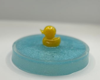 May include: A small yellow rubber ducky sits in the center of a light blue round tray with a glittery surface.