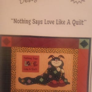 May include: A quilt pattern by Jane Wilson Designs, featuring a cartoon bunny wrapped in a quilt. The quilt has a central image with the words "Nothing Says Love Like A Quilt". The pattern includes a JW-79 pattern number.