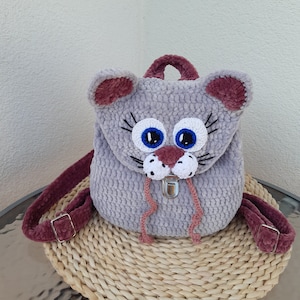 May include: A gray crocheted cat backpack with pink ears, blue eyes, and a white nose. The backpack has a silver buckle and pink straps.