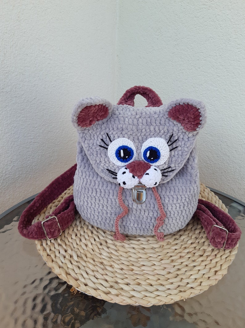 May include: A gray crocheted cat backpack with pink ears, blue eyes, and a white nose. The backpack has a silver buckle and pink straps.