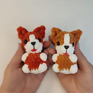 May include: Two handmade crochet corgi dog toys. The corgis are orange and white with black eyes and red mouths. The toys are held in two hands against a white background. The corgis have large ears and small paws.