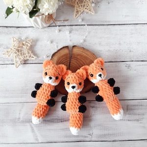 Op de afbeelding: Drie gehaakte vos knuffels met oranje vacht, zwarte ogen en zwarte neuzen. De vossen zitten op een houten oppervlak.