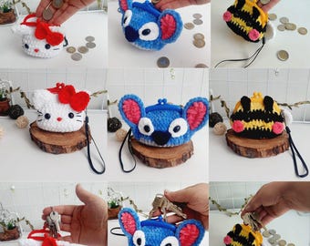Könnte beinhalten: Eine Auswahl handgefertigter gehäkelter Münztaschen mit Charakterdesigns. Die Taschen umfassen eine weiße Katze mit roter Schleife, einen blauen Alien mit rosa Ohren und eine gelb-schwarze Biene. Jede Tasche hat eine schwarze Handschlaufe.