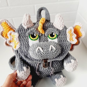 May include: A gray crocheted dragon backpack with yellow and pink accents. The backpack has a zipper closure and a small handle on top.
