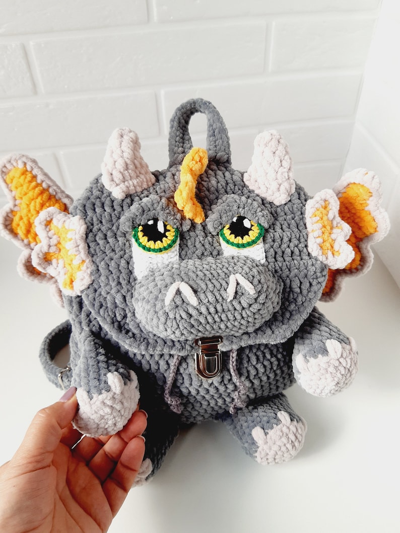 May include: A gray crocheted dragon backpack with yellow and pink accents. The backpack has a zipper closure and a small handle on top.