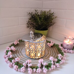 K&ouml;nnte beinhalten: Ein silberner Kerzenhalter aus Metall mit einem dekorativen Design und einem Glaseinsatz. Der Kerzenhalter steht auf einem geh&auml;kelten Untersetzer mit rosa und gr&uuml;nen Blumen. Der Untersetzer befindet sich auf einer wei&szlig;en Oberfl&auml;che.