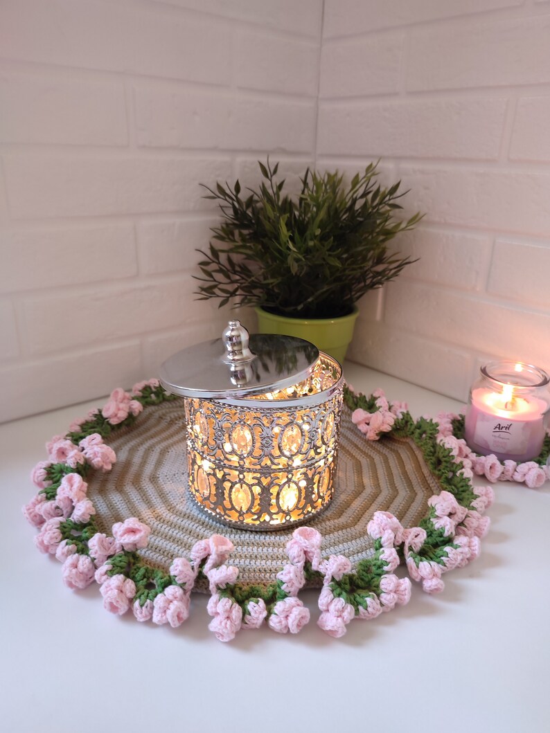 K&ouml;nnte beinhalten: Ein silberner Kerzenhalter aus Metall mit einem dekorativen Design und einem Glaseinsatz. Der Kerzenhalter steht auf einem geh&auml;kelten Untersetzer mit rosa und gr&uuml;nen Blumen. Der Untersetzer befindet sich auf einer wei&szlig;en Oberfl&auml;che.