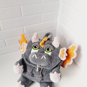 May include: Gray crocheted dragon backpack with orange wings and pink accents. The backpack has a zipper closure and green eyes.