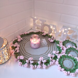 K&ouml;nnte beinhalten: Ein Set geh&auml;kelter Tischsets mit einem rosa-gr&uuml;nen Blumendesign. Die Tischsets sind rund und haben eine hellgraue Mitte. Eine Kerze steht auf einem der Tischsets. Das Wort "HOME" ist in wei&szlig;en Buchstaben mit Lichtern dahinter geschrieben.