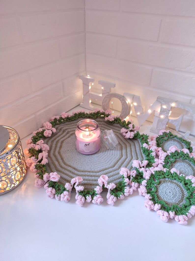 K&ouml;nnte beinhalten: Ein Set geh&auml;kelter Tischsets mit einem rosa-gr&uuml;nen Blumendesign. Die Tischsets sind rund und haben eine hellgraue Mitte. Eine Kerze steht auf einem der Tischsets. Das Wort "HOME" ist in wei&szlig;en Buchstaben mit Lichtern dahinter geschrieben.