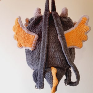 May include: A gray crocheted backpack with orange wings and a tail. The backpack has adjustable straps.