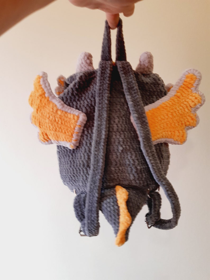 May include: A gray crocheted backpack with orange wings and a tail. The backpack has adjustable straps.