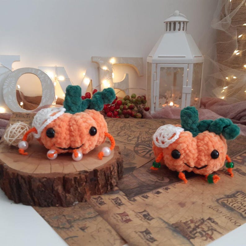 May include: Two crocheted orange pumpkin plushies with green stems and white crocheted accents. The pumpkins are sitting on a wooden surface with a map in the background.