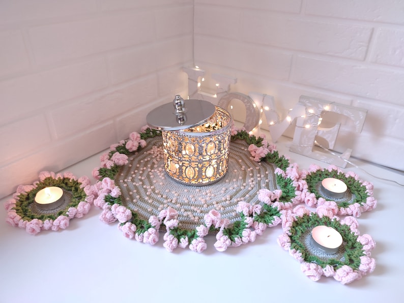 K&ouml;nnte beinhalten: Eine silberne dekorative Metallbox mit Deckel, umgeben von drei geh&auml;kelten Untersetzern mit rosa Bl&uuml;ten und gr&uuml;nen Bl&auml;ttern. Die Untersetzer befinden sich auf einer wei&szlig;en Oberfl&auml;che mit einem wei&szlig;en Holzschild, auf dem "LOVE" mit Lichtern steht.