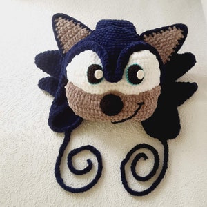 Könnte beinhalten: Eine blaue und braune Häkelmütze in Form von Sonic the Hedgehog. Die Mütze hat ein weißes Gesicht mit schwarzen Augen und einer schwarzen Nase. Die Mütze hat eine lange blaue Schnur, die zu einem Knoten gebunden ist.