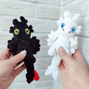 May include: Two handmade crochet dragon plushies. One is black with yellow eyes and a red tail fin. The other is white with blue eyes. Both are held against a white brick background.