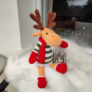 May include: A crocheted reindeer toy wearing a green and white striped sweater, a red scarf, and red mittens. The reindeer is sitting on a white fluffy surface.
