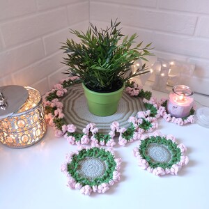 K&ouml;nnte beinhalten: Ein Set geh&auml;kelter Untersetzer und eine gr&ouml;&szlig;ere geh&auml;kelte Tischdecke in Gr&uuml;n und Pink mit einer kleinen Topfpflanze in der Mitte.
