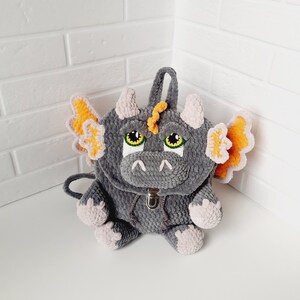 May include: Gray crocheted dragon backpack with orange wings and pink feet. The backpack has a zipper closure and a small handle on the top.