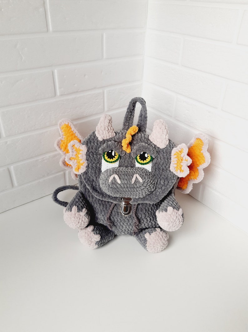 May include: Gray crocheted dragon backpack with orange wings and pink feet. The backpack has a zipper closure and a small handle on the top.