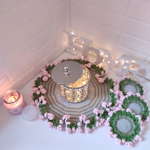 K&ouml;nnte beinhalten: Ein Set geh&auml;kelter Untersetzer und ein gr&ouml;&szlig;erer geh&auml;kelter Platzdeckchen mit rosa und gr&uuml;nen Akzenten. Das Platzdeckchen hat einen silbernen Metallbeh&auml;lter mit Deckel in der Mitte. Das Wort "LOVE" ist in wei&szlig;en Buchstaben mit Lichtern an einer wei&szlig;en Backsteinwand im Hintergrund geschrieben.