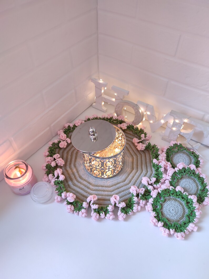 K&ouml;nnte beinhalten: Ein Set geh&auml;kelter Untersetzer und ein gr&ouml;&szlig;erer geh&auml;kelter Platzdeckchen mit rosa und gr&uuml;nen Akzenten. Das Platzdeckchen hat einen silbernen Metallbeh&auml;lter mit Deckel in der Mitte. Das Wort "LOVE" ist in wei&szlig;en Buchstaben mit Lichtern an einer wei&szlig;en Backsteinwand im Hintergrund geschrieben.