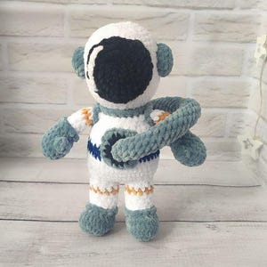 May include: A crocheted astronaut toy with a white body, blue arms and legs, and a black face. The astronaut is holding a blue arm in front of its body.