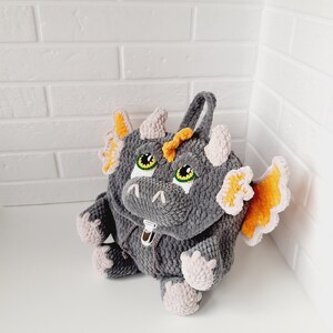 May include: A gray crocheted dragon backpack with orange wings and green eyes. The backpack has a zipper closure and a pink crocheted lining.