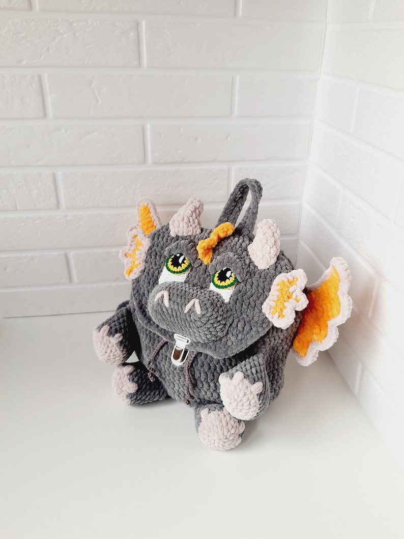 May include: A gray crocheted dragon backpack with orange wings and green eyes. The backpack has a zipper closure and a pink crocheted lining.