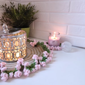 K&ouml;nnte beinhalten: Eine geh&auml;kelte Unterlage mit rosa und gr&uuml;nen Blumen umgibt einen silbernen Metallbeh&auml;lter mit Deckel und eine rosa Kerze in einem Glasgef&auml;&szlig;. Die Kerze ist angez&uuml;ndet und der Beh&auml;lter hat ein Licht im Inneren.