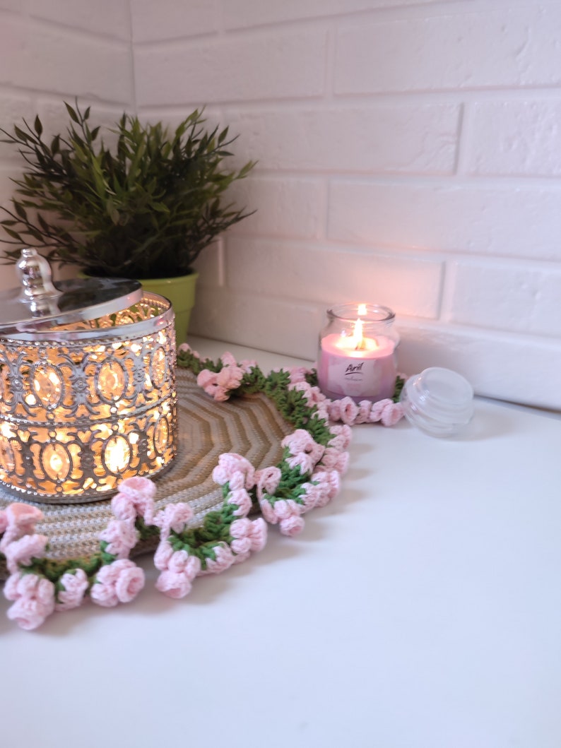 K&ouml;nnte beinhalten: Eine geh&auml;kelte Unterlage mit rosa und gr&uuml;nen Blumen umgibt einen silbernen Metallbeh&auml;lter mit Deckel und eine rosa Kerze in einem Glasgef&auml;&szlig;. Die Kerze ist angez&uuml;ndet und der Beh&auml;lter hat ein Licht im Inneren.