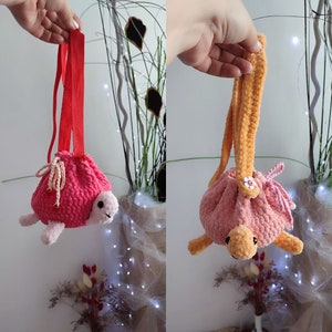 May include: Two crocheted turtle-shaped drawstring bags with pink fabric and different coloured straps. One bag has a red strap and the other has an orange strap.