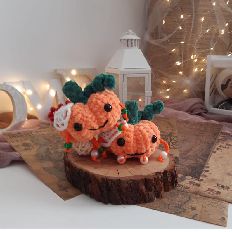 May include: Two crocheted pumpkin plushies with green stems and smiling faces. The pumpkins are orange and have white accents. They are sitting on a wooden slice.