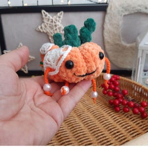 May include: A crocheted pumpkin-shaped plush toy with a green stem and white crocheted details. The toy has a smiling face with black eyes and a white mouth. The toy is holding white pearl beads in its arms.