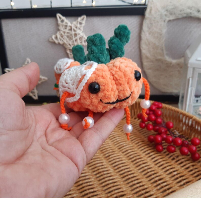 May include: A crocheted pumpkin-shaped plush toy with a green stem and white crocheted details. The toy has a smiling face with black eyes and a white mouth. The toy is holding white pearl beads in its arms.