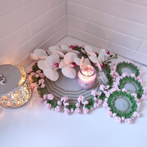K&ouml;nnte beinhalten: Ein Set geh&auml;kelter Untersetzer mit einem rosa-gr&uuml;nen Blumendesign. Die Untersetzer sind aus hellgrauem Garn gefertigt und haben einen gewellten Rand. Eine rosa Kerze steht auf einem gr&ouml;&szlig;eren geh&auml;kelten Untersetzer mit einem wei&szlig;en Orchideen-Blumenarrangement.