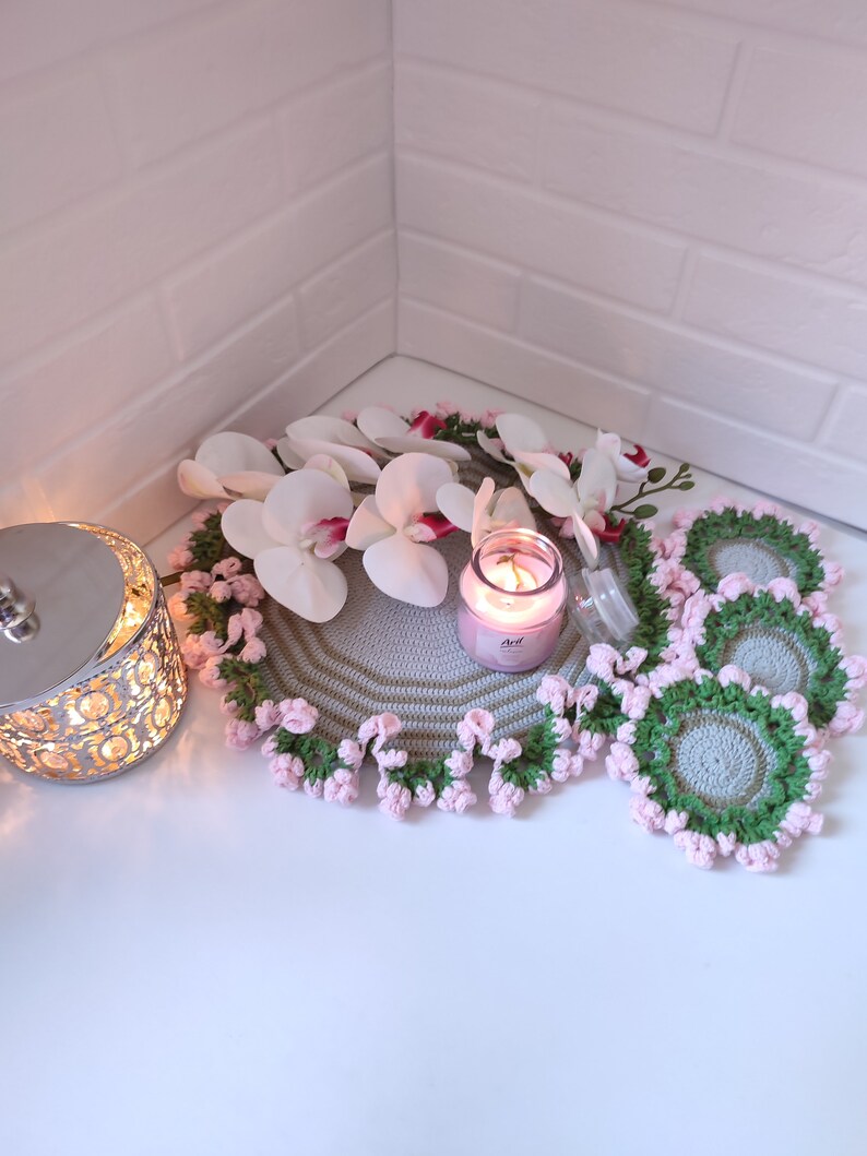 K&ouml;nnte beinhalten: Ein Set geh&auml;kelter Untersetzer mit einem rosa-gr&uuml;nen Blumendesign. Die Untersetzer sind aus hellgrauem Garn gefertigt und haben einen gewellten Rand. Eine rosa Kerze steht auf einem gr&ouml;&szlig;eren geh&auml;kelten Untersetzer mit einem wei&szlig;en Orchideen-Blumenarrangement.